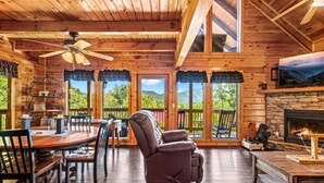 A cozy cabin interior with a dining area, a fireplace, and large windows offering scenic views. - 2BR w/Hot Tub*Fireplace, MTN View, Community Pool! (Sevierville)