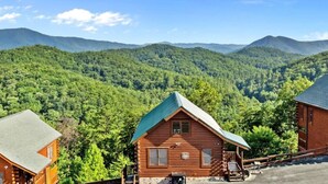 A cabin with a stone foundation and a green roof, surrounded by lush forest and mountains. - 2BR Cabin w/MTN Views*Hot Tub*Games*Community Pool (Sevierville)