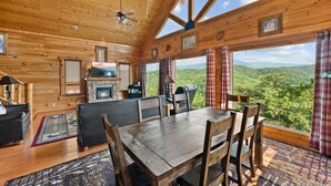 A wooden cabin interior with a dining table, chairs, a fireplace, a television, and large windows offering a view of the forest. - 2BR Cabin w/MTN Views*Hot Tub*Games*Community Pool (Sevierville)