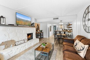 A modern living room with a fireplace, a dining area, and a kitchen visible in the background.