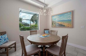 Un coin repas avec une table ronde en bois, six chaises, un lustre et deux grandes fenêtres donnant sur un paysage verdoyant et une plage.