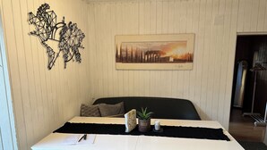 Un coin repas comprenant une table blanche, deux chaises blanches, un banc noir, un élément décoratif mural et un tableau représentant un paysage urbain, encadré.