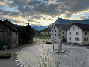 A roundabout with a central sculpture, surrounded by buildings with traditional architecture, a clear sky, and mountains in the background. - Central but quietly located, lovely and well furnished flat in Balzers. (Balzers)