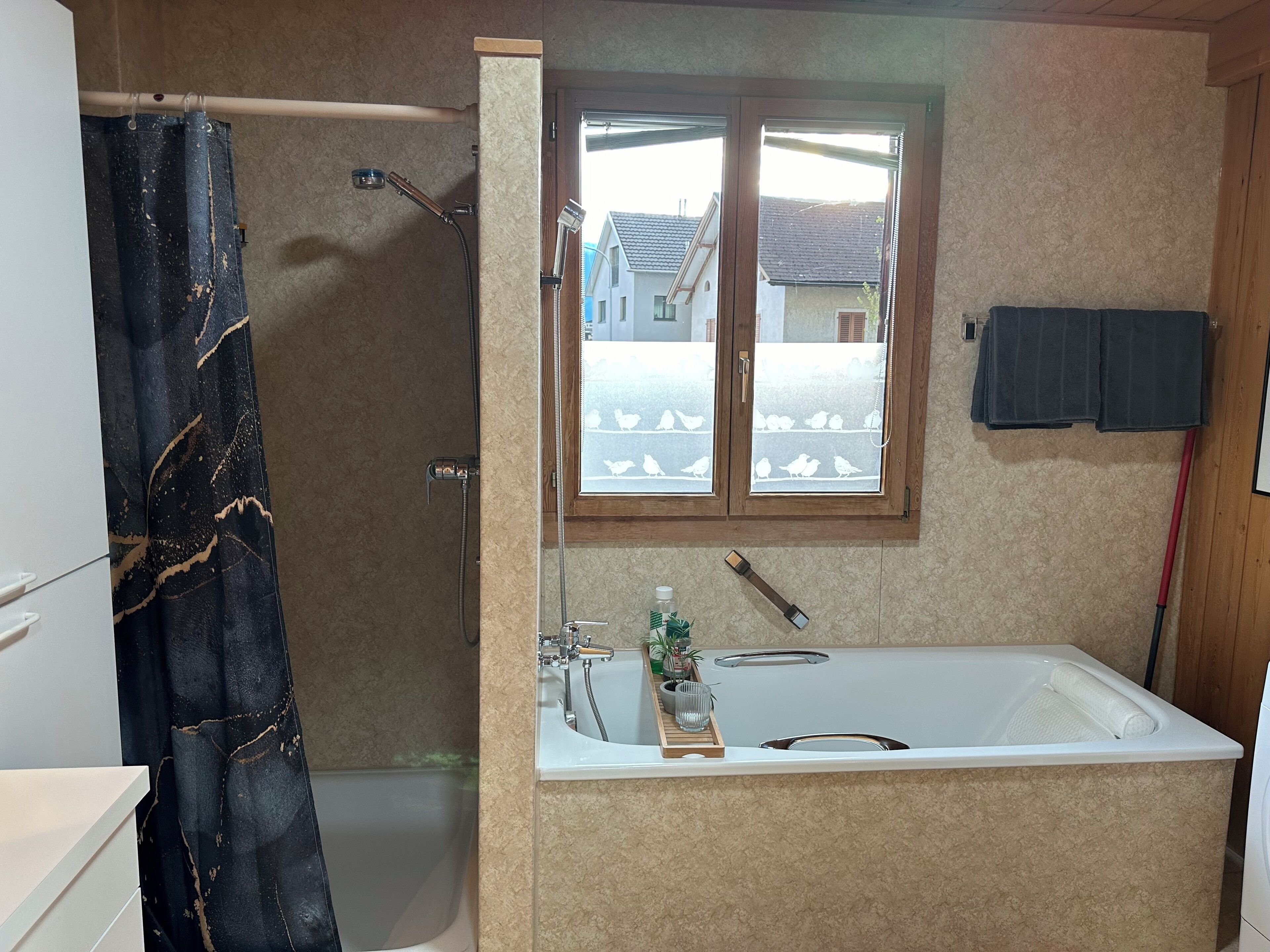 Een badkamer met een bad, een douche, een raam met uitzicht op huizen en een handdoekrek.