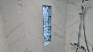 Un bany modern amb parets de marbre, una dutxa amb plat de roba de mà i un moble prestatge de vidre.