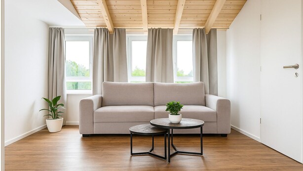 A modern living room with a grey sofa, a round table with a plant, and a wooden ceiling with exposed beams. - Lux Suites Leipzig Paradise Apartment (Leipzig)