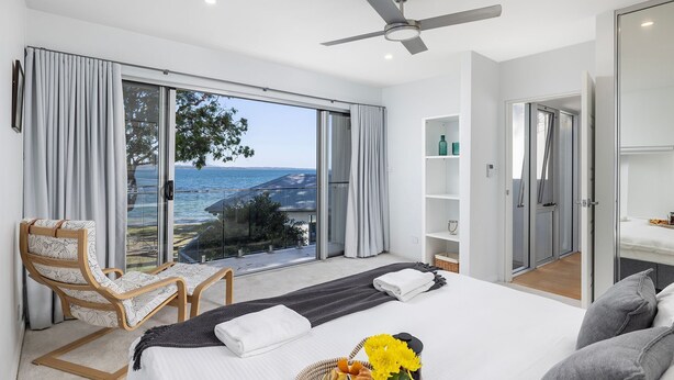 A modern bedroom with a large bed, a chair, a ceiling fan, and a view of the sea.
