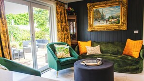 A modern living room with a green sofa, a green armchair, and a round ottoman. There is a painting on the wall and a glass door leading to an outdoor area. - Lore Haven - Your little slice of paradise (Robe)
