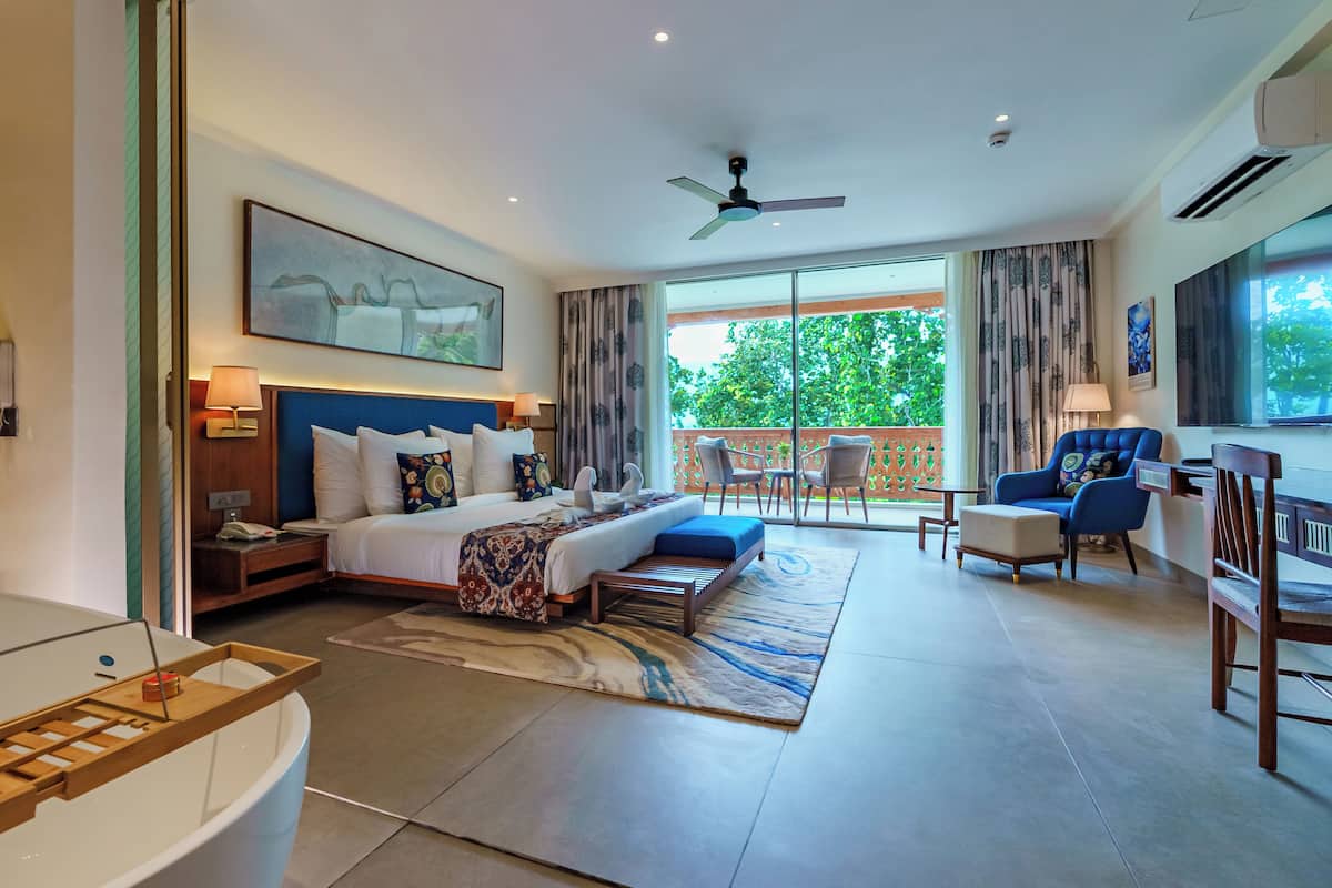 A modern hotel room with a large bed, a sitting area with a sofa and armchairs, a balcony with a view, and a bathroom with a bathtub.