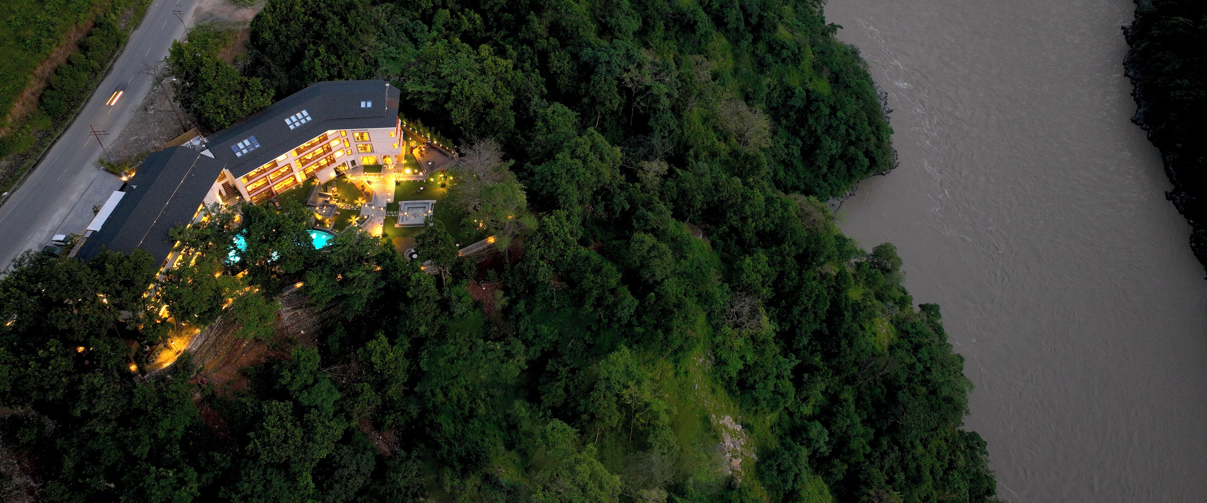 An aerial view of a hotel with a swimming pool, surrounded by dense forest and a river.