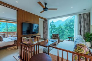 A modern hotel room with a large TV, wooden furniture, a balcony with a view of mountains, and a ceiling fan.