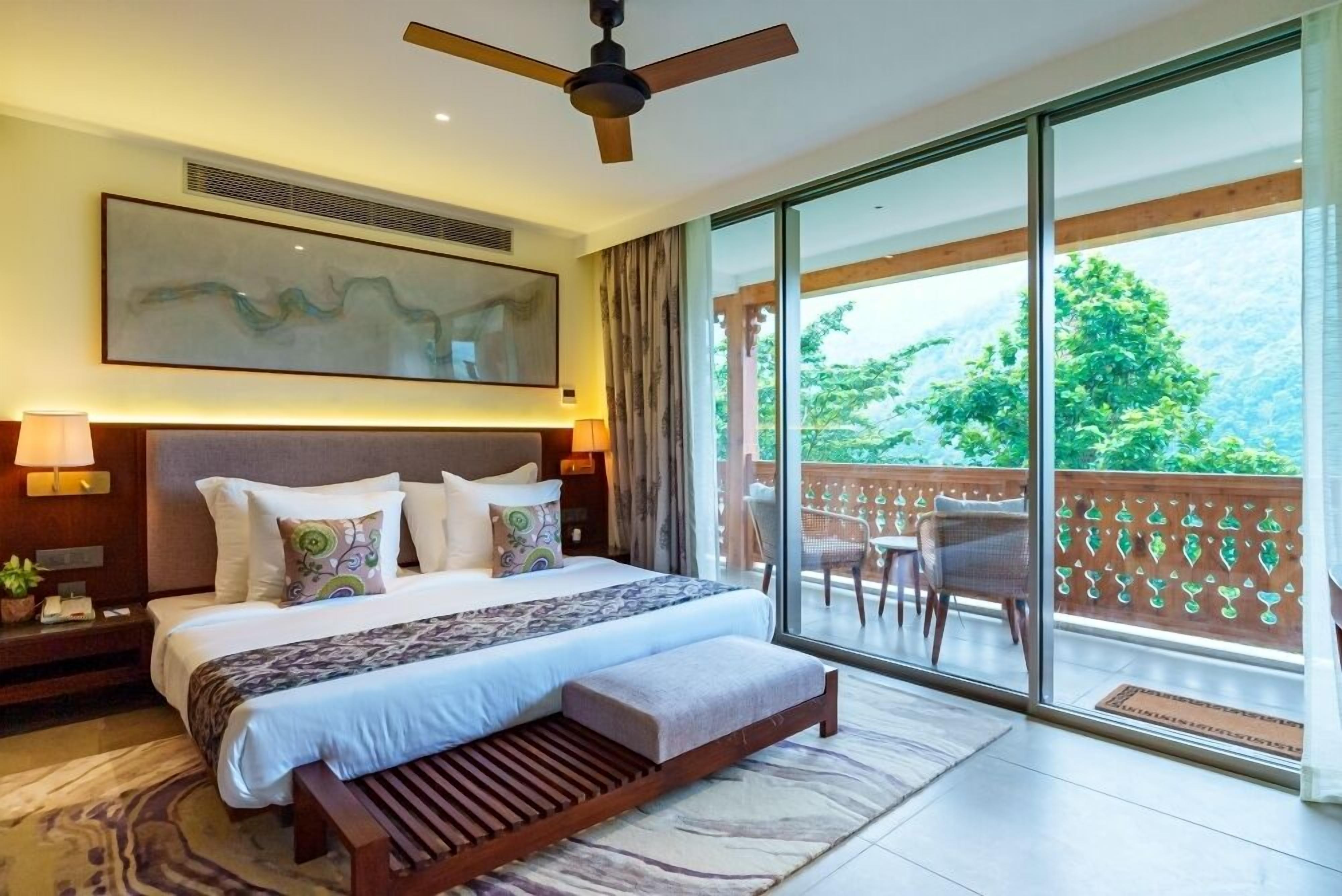 A modern hotel room with a large bed, a ceiling fan, a balcony with a view of greenery, and a large window.
