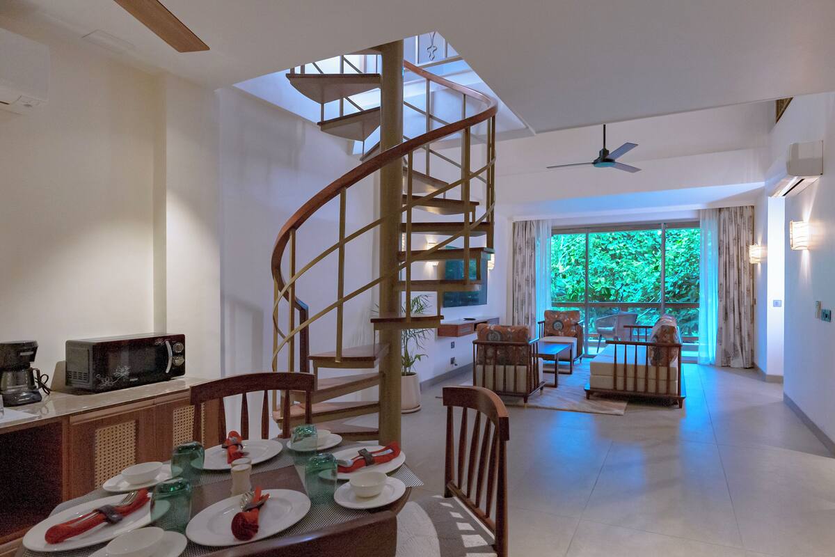 A dining area with a spiral staircase, a dining table set with plates and cutlery, and a view of a balcony with greenery.