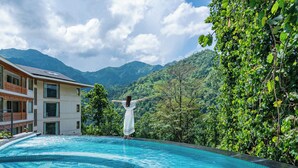 A swimming pool with a person standing in the water, surrounded by lush greenery and mountains in the background. - VILEEN Rishikesh - A Wellness Resort by Ganga Kinare (Rishikesh)