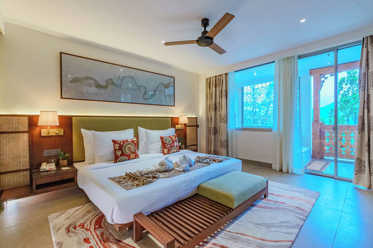 A spacious hotel room with a large bed, a ceiling fan, a large window with a view of greenery, and a wall-mounted artwork.