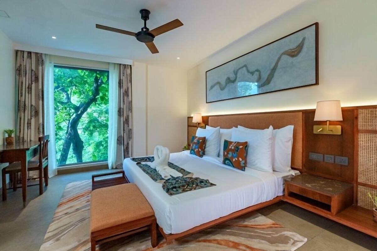 A hotel room with a large bed, a ceiling fan, a view of trees through the window, and a painting on the wall.