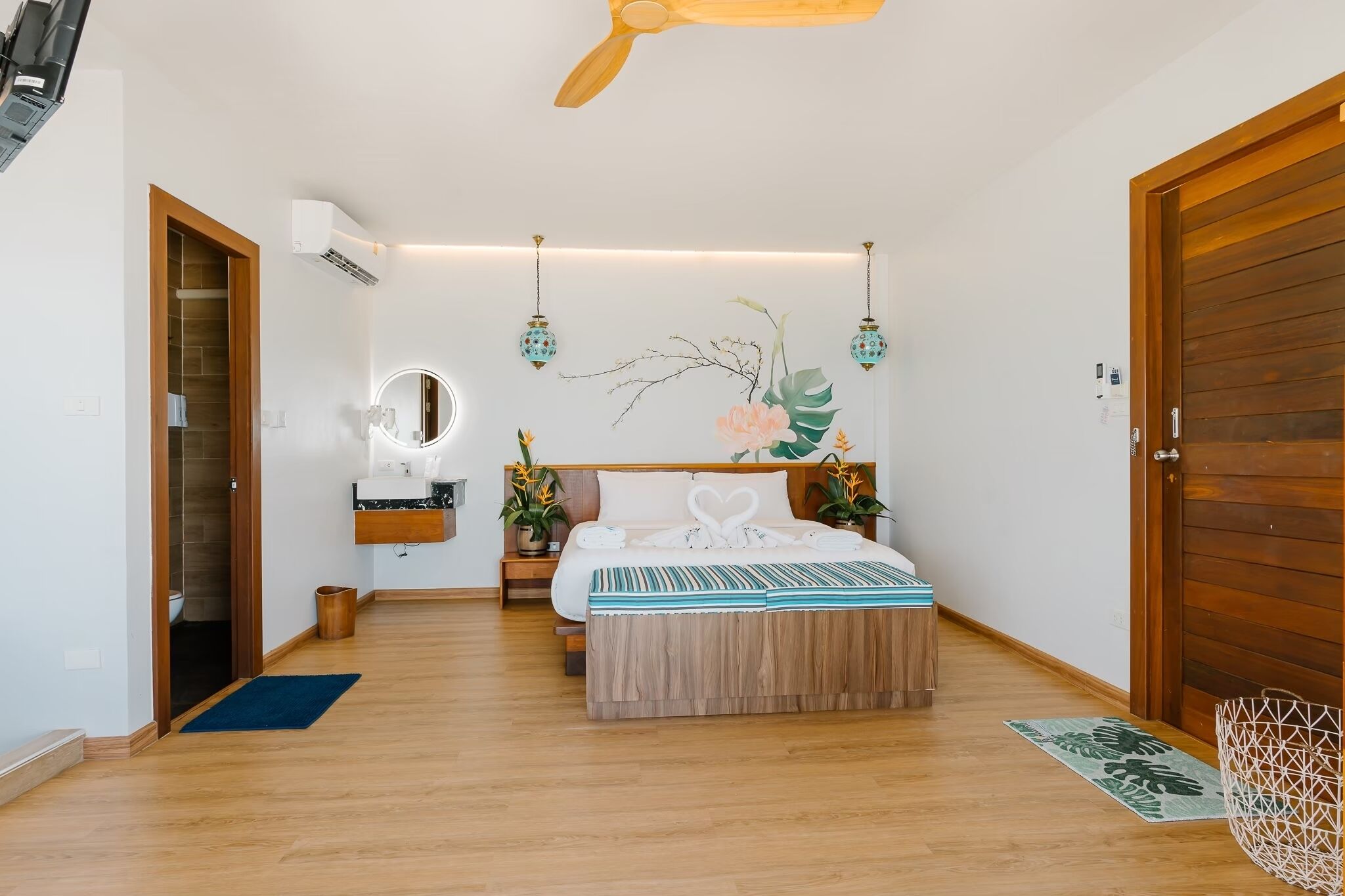 A modern bedroom with a wooden floor, a bed with a heart-shaped pillow, a bedside table, a mirror, and a wall-mounted air conditioner.