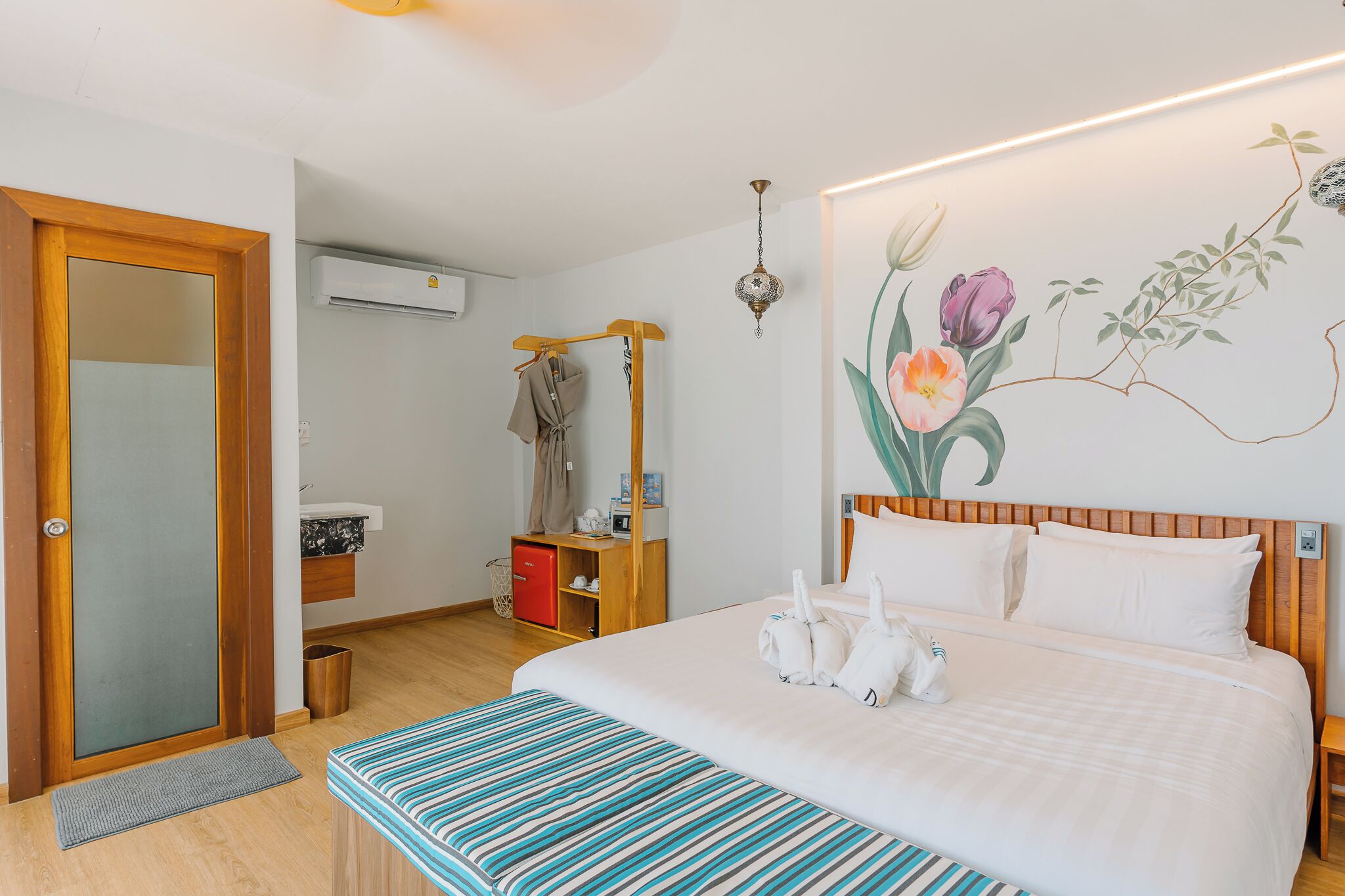 Una habitación de hotel con una cama, un cabecero de madera, un aire acondicionado de pared, un pequeño armario rojo, un banco y una pared con un mural floral.