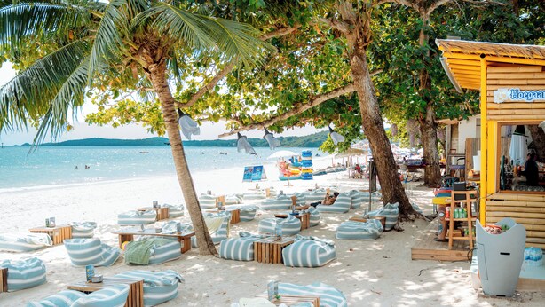 A beachside area with bean bag chairs and tables under trees, a wooden structure with a sign, and a view of the sea.