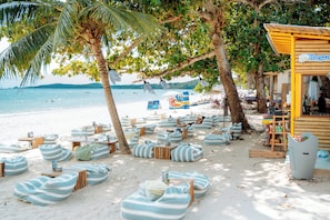A beachside area with bean bag chairs and tables under trees, a wooden structure with a sign, and a view of the sea.