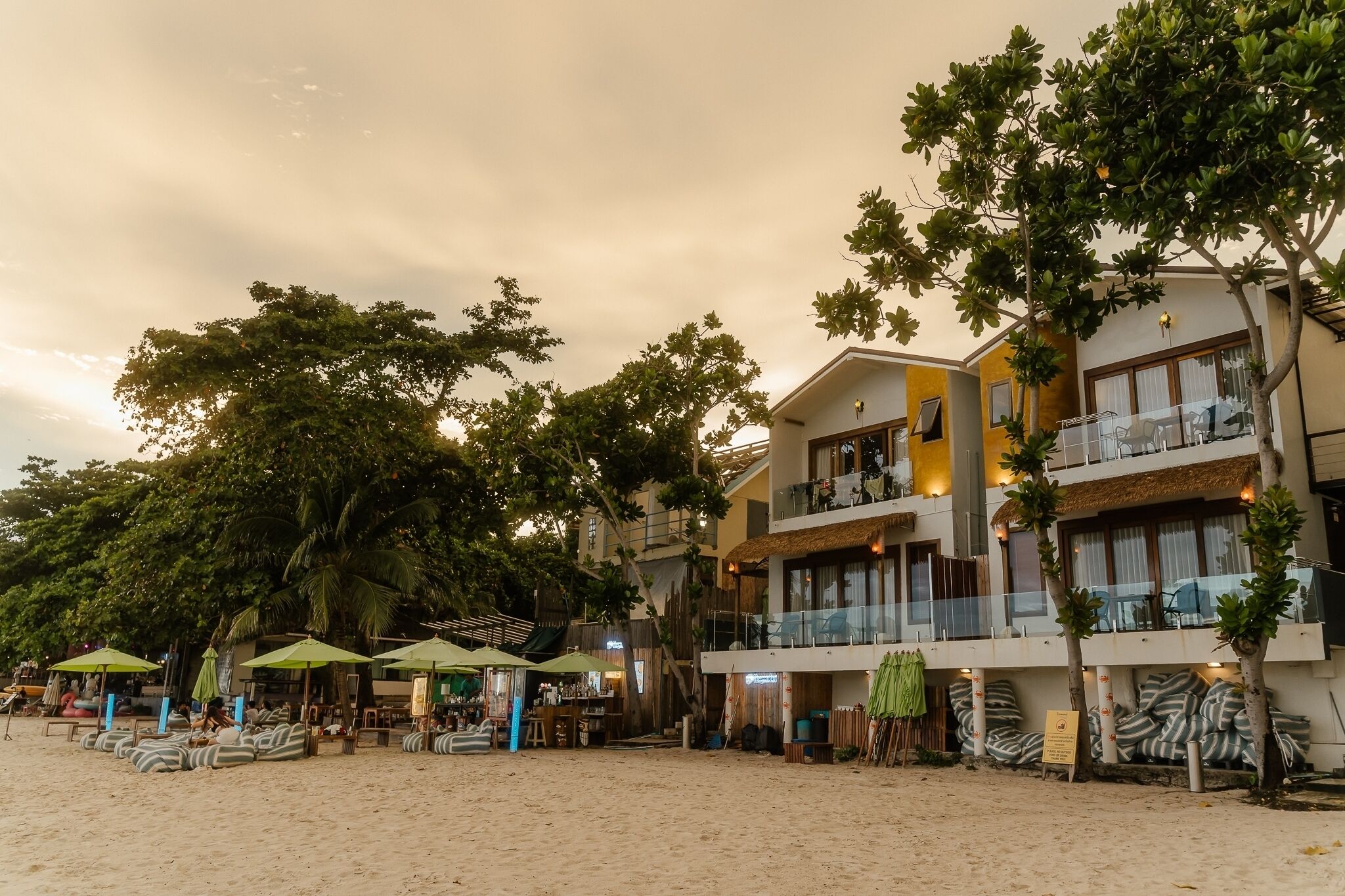 A beachfront hotel with multiple balconies, outdoor seating, and a thatched-roof structure.
