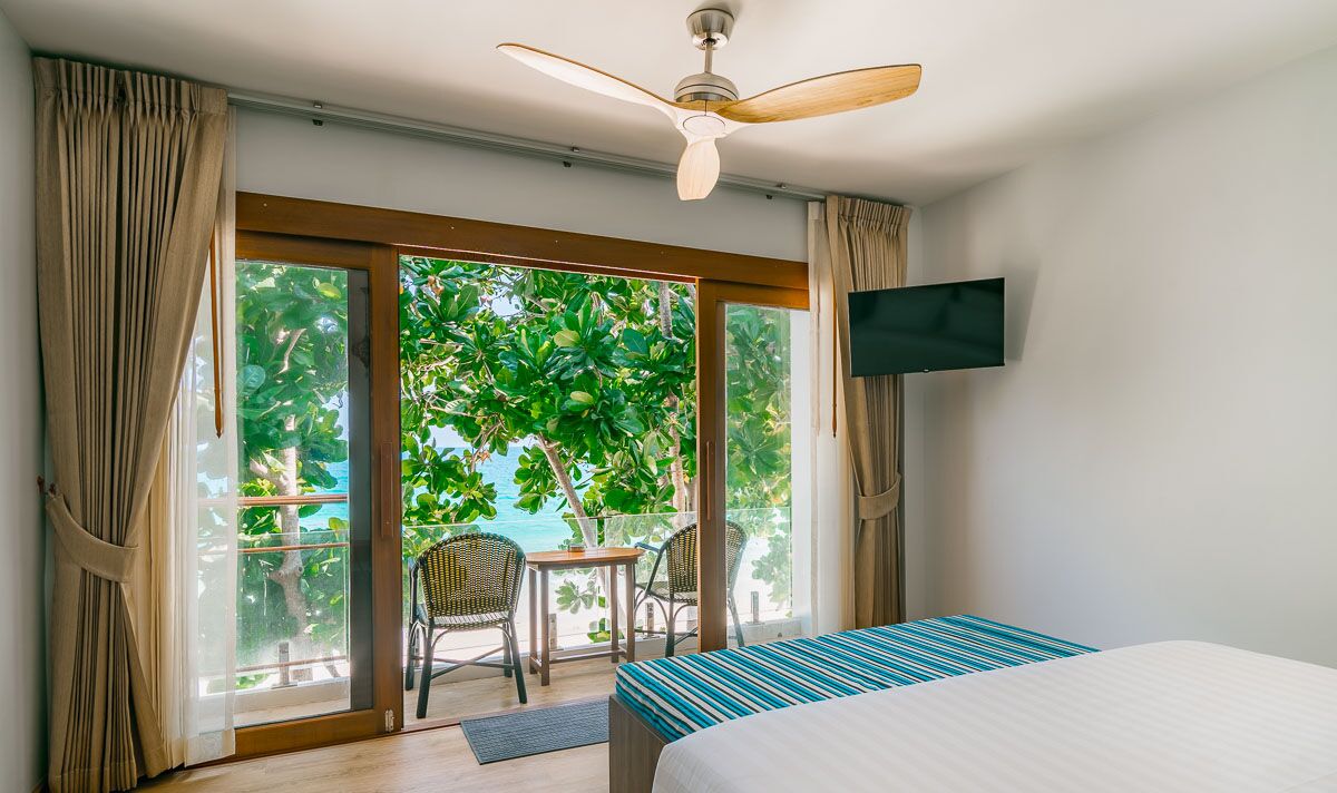 A hotel room with a bed, a ceiling fan, a TV mounted on the wall, and a balcony with a table and chairs.