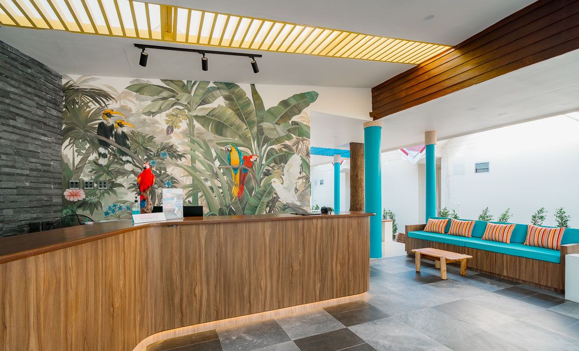 A reception area with a wooden reception desk, a large mural of parrots and foliage, a teal sofa, and a checkered floor.