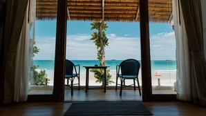 A room with a view of the beach, featuring a table and two chairs, a plant, and a thatched roof.