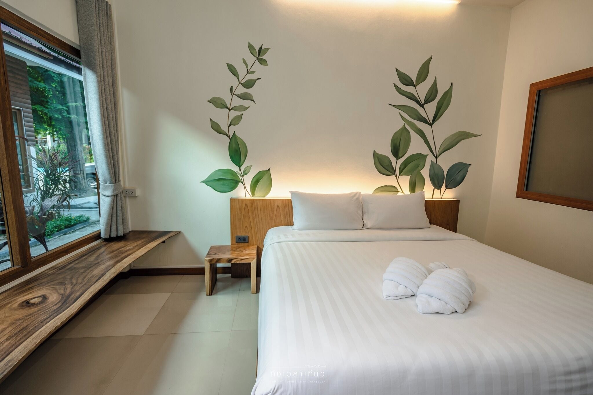 A neatly made bed with white linens and two pillows, a small wooden nightstand with a lamp, and two wall-mounted plants.