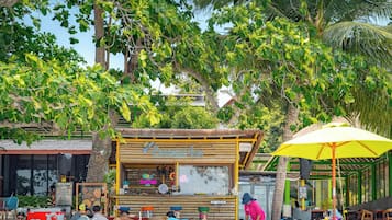 Un café al aire libre con techo de paja, rodeado de exuberante follaje y palmeras.