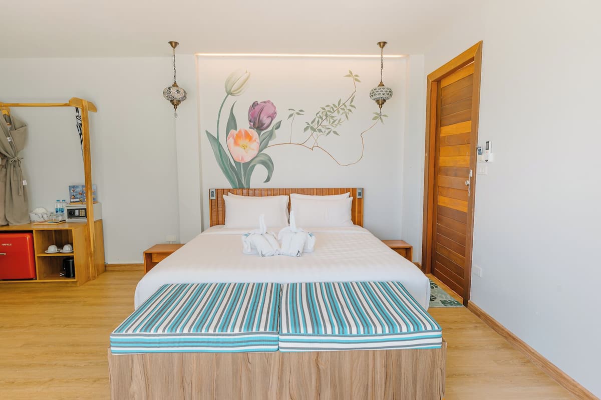 A neatly made bed with white linens and a striped blanket, a wooden nightstand with a lamp, a red cabinet, and a wall-mounted mirror.