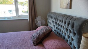 A bedroom with a tufted headboard, a bed with pink bedding, a patterned pillow, a standing fan, and a window with curtains. - Leeds - Apartment - Stay 3- Parking - Garden (Leeds)