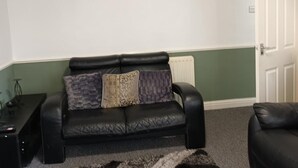 A living room with a black leather sofa, two patterned cushions, a small black side table, and a radiator. - Leeds - Apartment - Stay 3- Parking - Garden (Leeds)