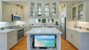 Een moderne keuken met witte kasten, een ingebouwde oven, een magnetron, en een groot kookeiland met een televisiescherm waarop een strand te zien is.
