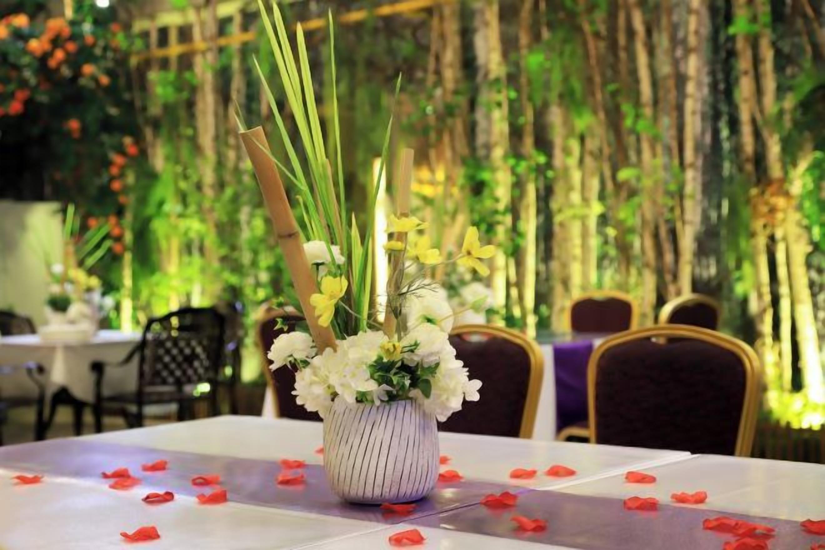 A table with a vase of flowers and red rose petals on a tablecloth.