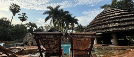 Zona de piscina con dos sillones de madera mirando a la piscina, rodeada de vegetación tropical y una estructura de techo de paja.