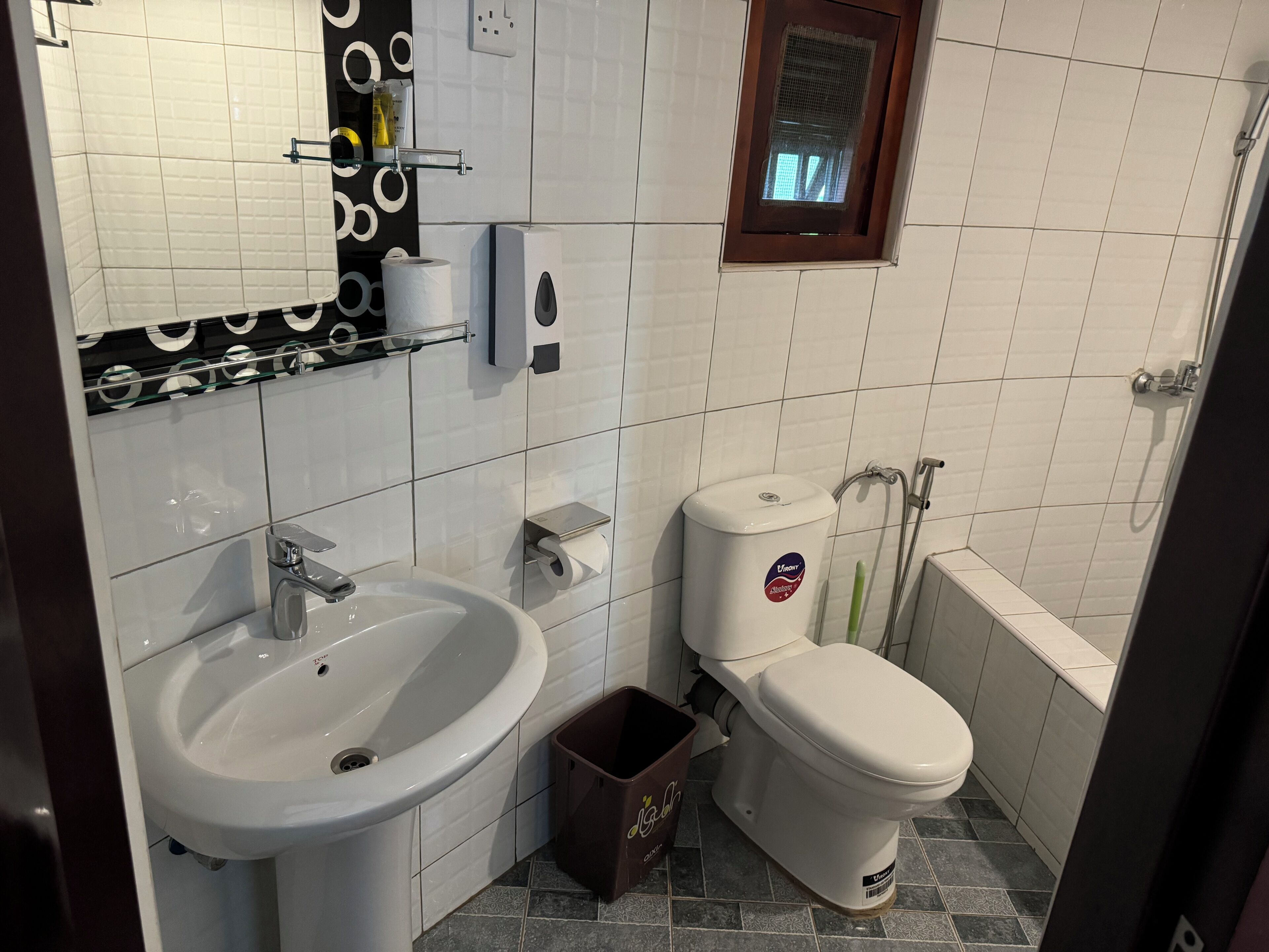 A bathroom with a white sink, toilet, and shower. There is a small window above the toilet and a towel rack above the sink.