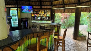 Een terras met een rieten dak, stenen vloer en verschillende barkrukken en stoelen.