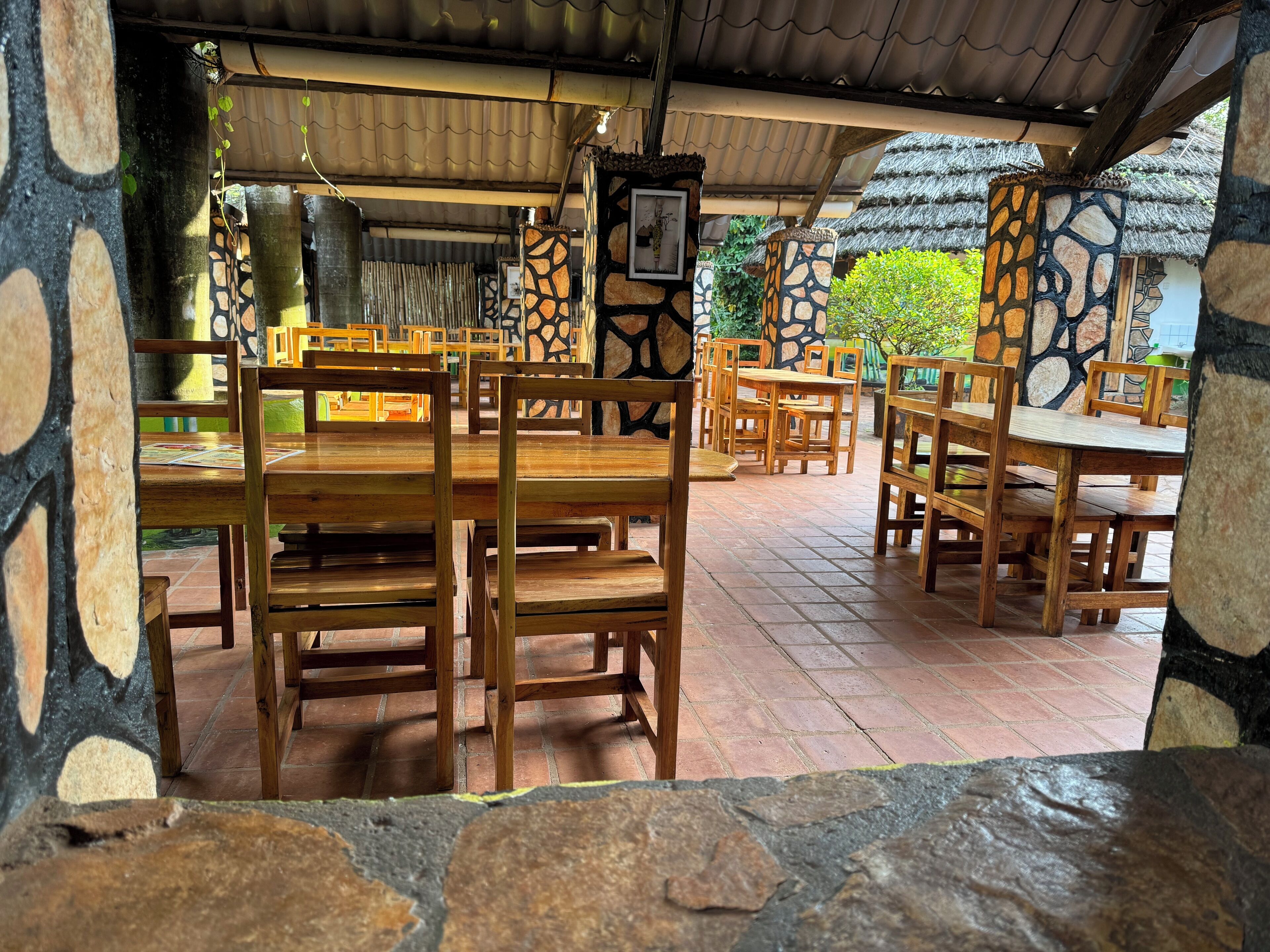 A covered outdoor dining area with wooden tables and chairs, stone walls, and a thatched roof.