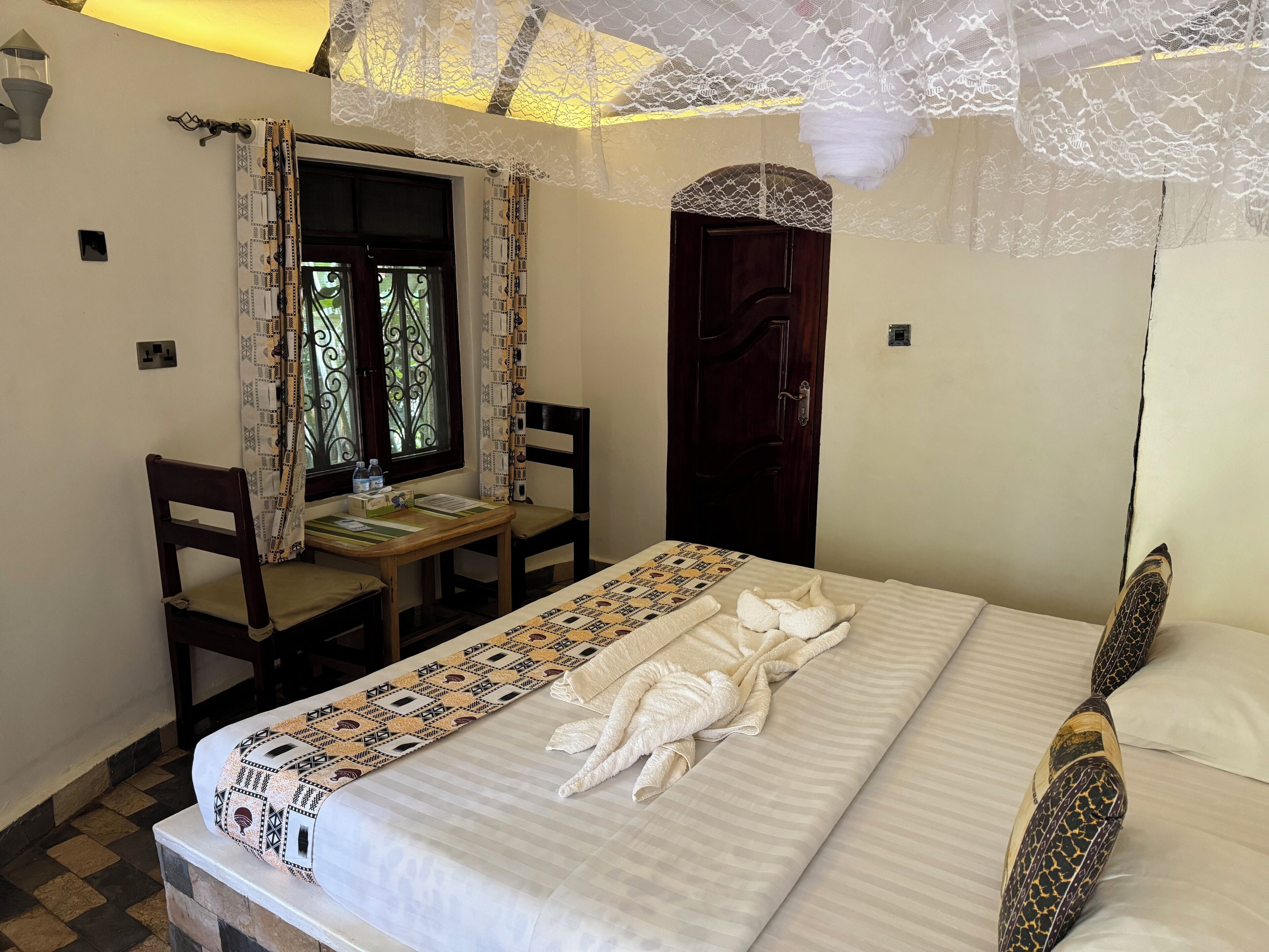 A neatly made bed with a patterned bedspread, a wooden desk with a chair, a window with decorative curtains, and a door with a lace curtain.