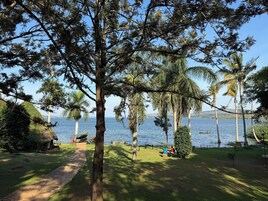 Un parque tranquilo junto al lago con una cabaña de techo de paja, un sendero pavimentado y una zona verde donde hay gente sentada en bancos.