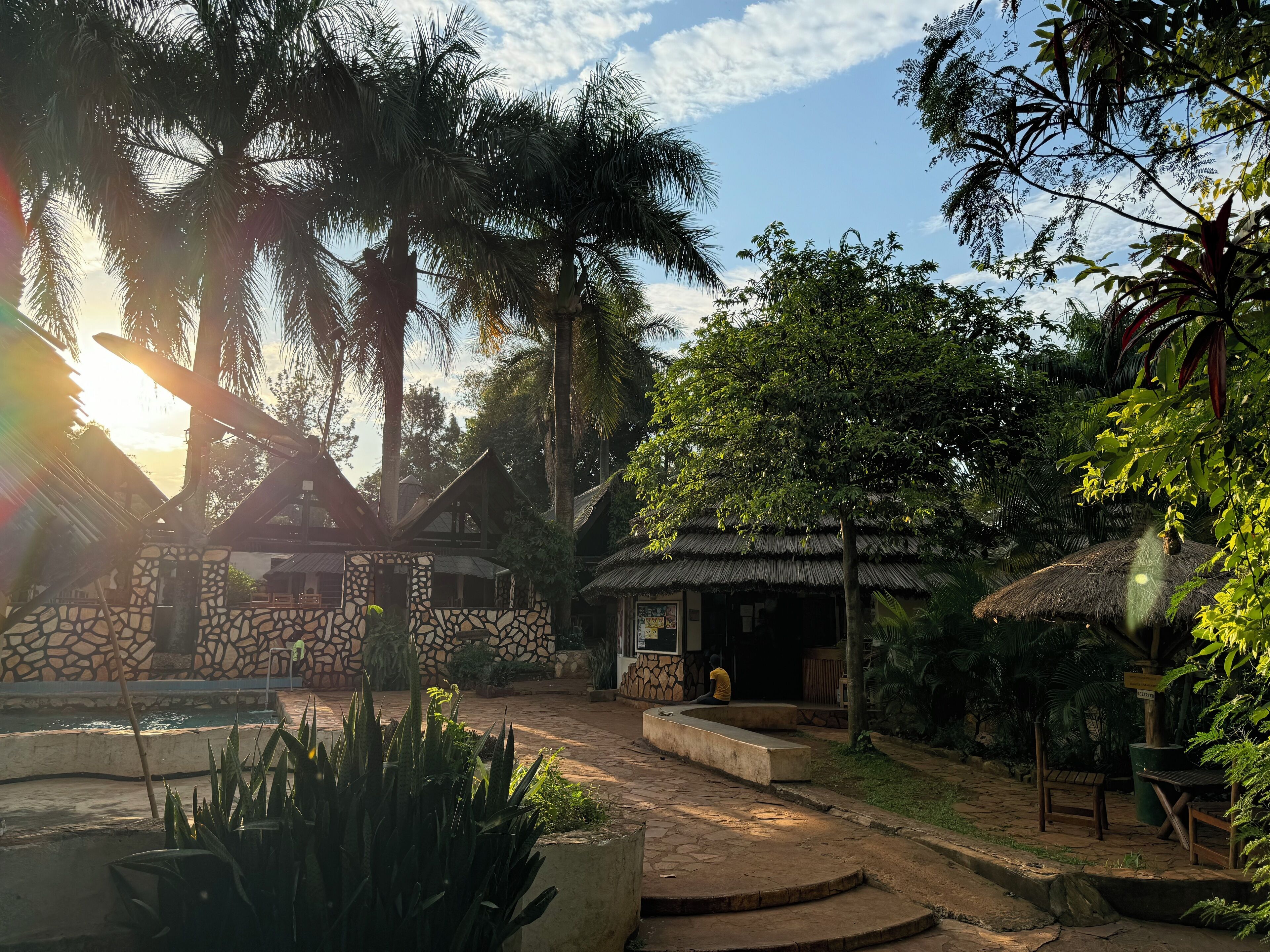 Un patio con muros de piedra, una construcción con techo de paja y vegetación tropical.