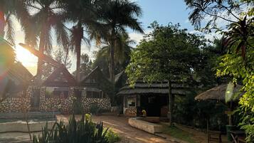 Un patio con muros de piedra, una construcción con techo de paja y vegetación tropical.
