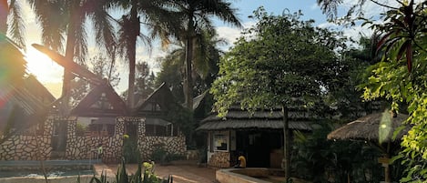 Un patio con muros de piedra, una construcción con techo de paja y vegetación tropical.