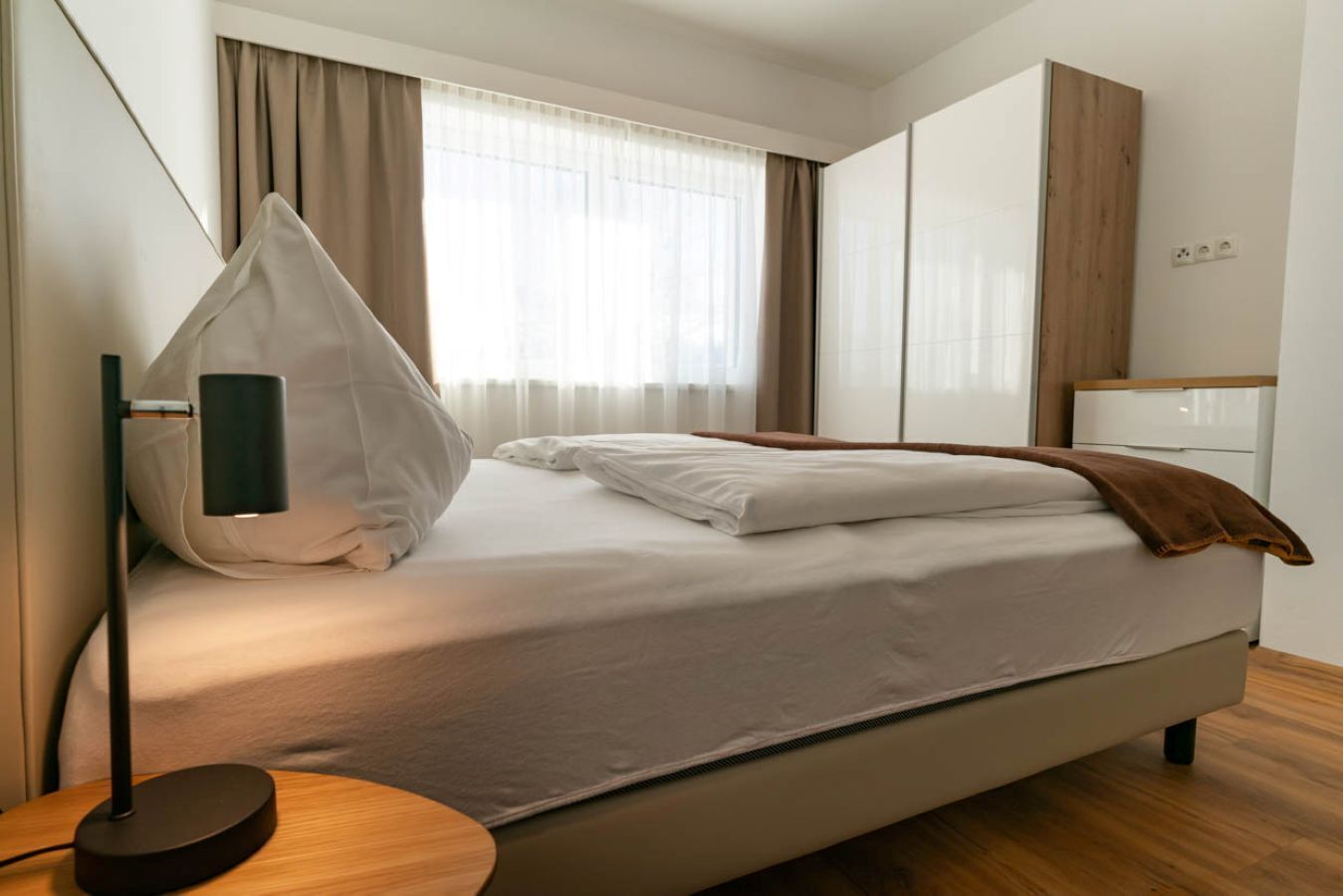 A modern hotel room with a bed, a bedside table, a lamp, and a wardrobe.