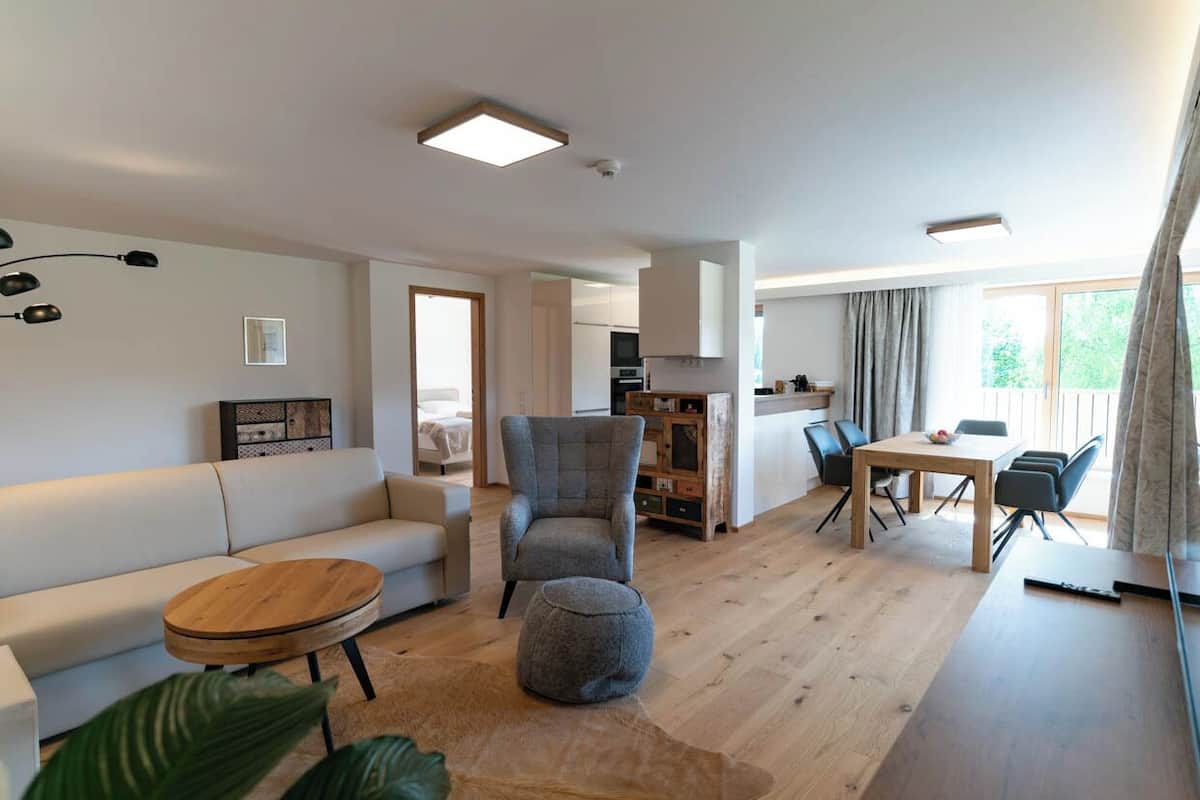 Un salon moderne avec un canapé, un pouf et une table basse ronde. On y trouve également un coin repas avec une table et des chaises. La pièce est dotée d’un parquet et d’une grande fenêtre agrémentée de rideaux.