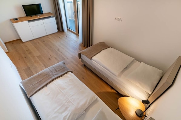 A compact room with two beds, a small desk, a TV, and a window with curtains. - Waldrand Apartments (Ramsau am Dachstein)