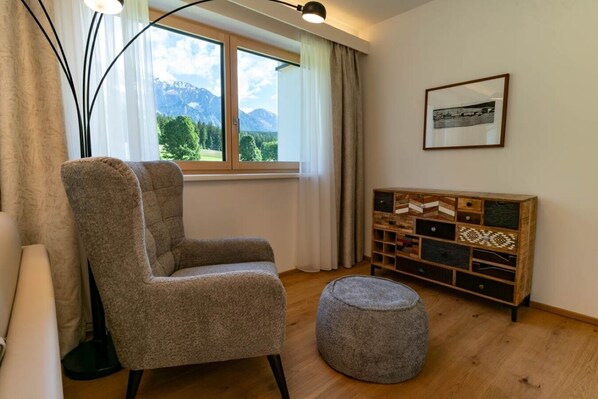 A living room with a large window, a chair, a ottoman, and a wooden sideboard. - Waldrand Apartments (Ramsau am Dachstein)