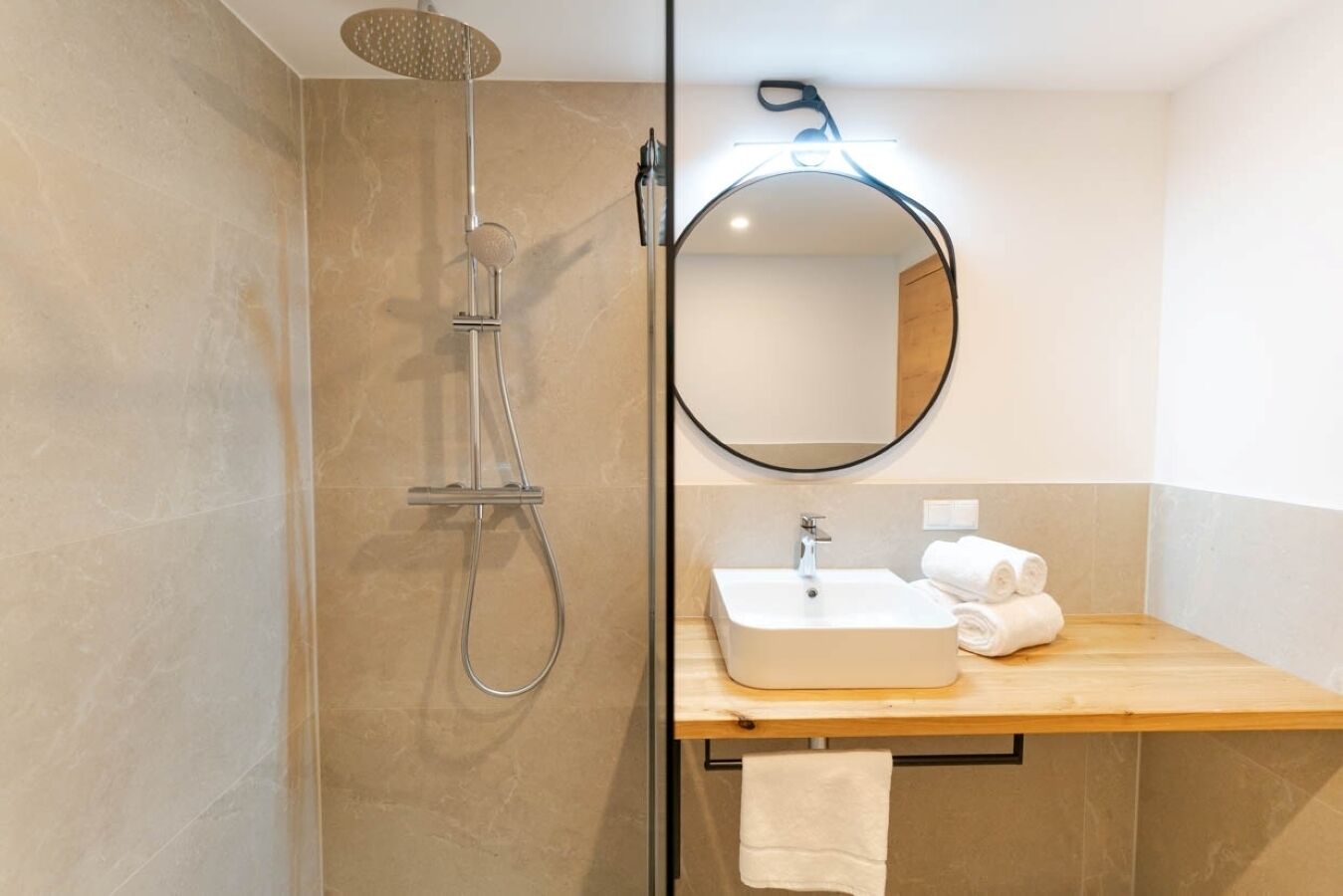 Ein modernes Badezimmer mit einer Duschkabine aus Glas, einem runden Spiegel und einem Holzwaschtisch mit Waschbecken und Handtüchern.