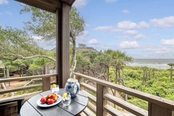 A balcony with a table set for a meal, overlooking a beach and a forested area.